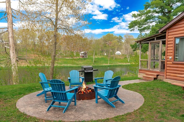 Blue Adirondack chairs around fire pit on patio, log cabin and pond with wooded grounds beyond